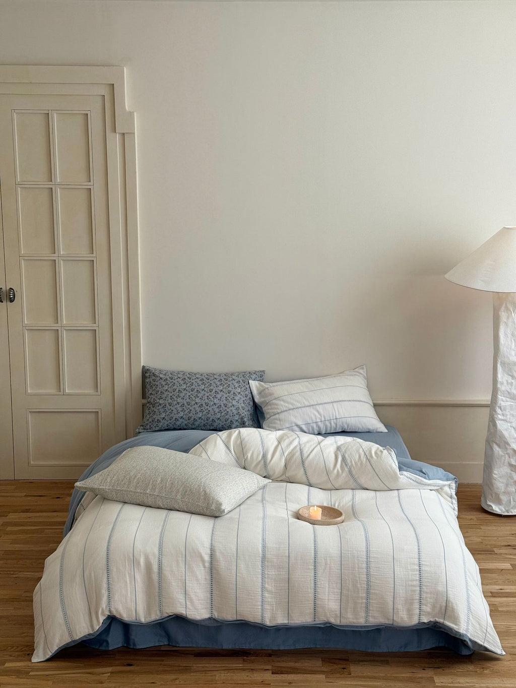 Bed with striped bedding and pillows in a room with a lamp and door.