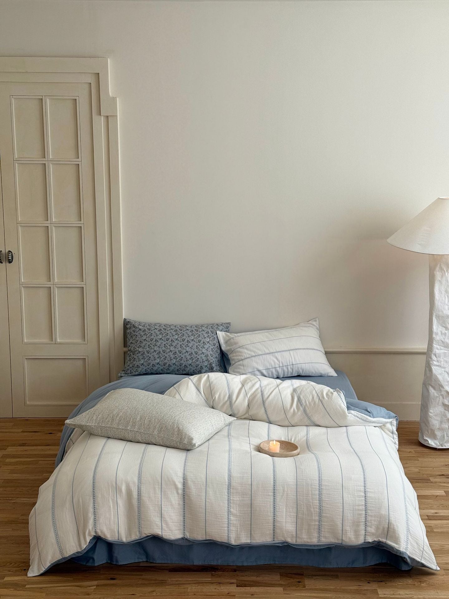 Bed with striped bedding and pillows in a room with a lamp and door.