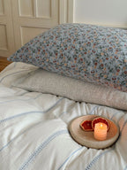 Floral pillow on a bed with a candle and dried fruit on a wooden coaster.