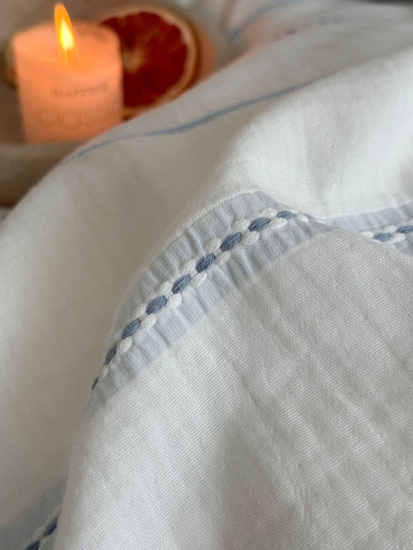 Close-up of a light blue fabric with decorative stitching, a lit candle, and a sliced grapefruit in the background.