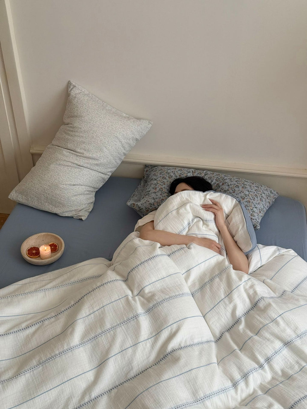 Person lying in bed under a striped duvet with pillows and a small bowl on the nightstand.