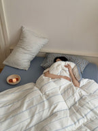 Person lying in bed under a striped duvet with pillows and a small bowl on the nightstand.