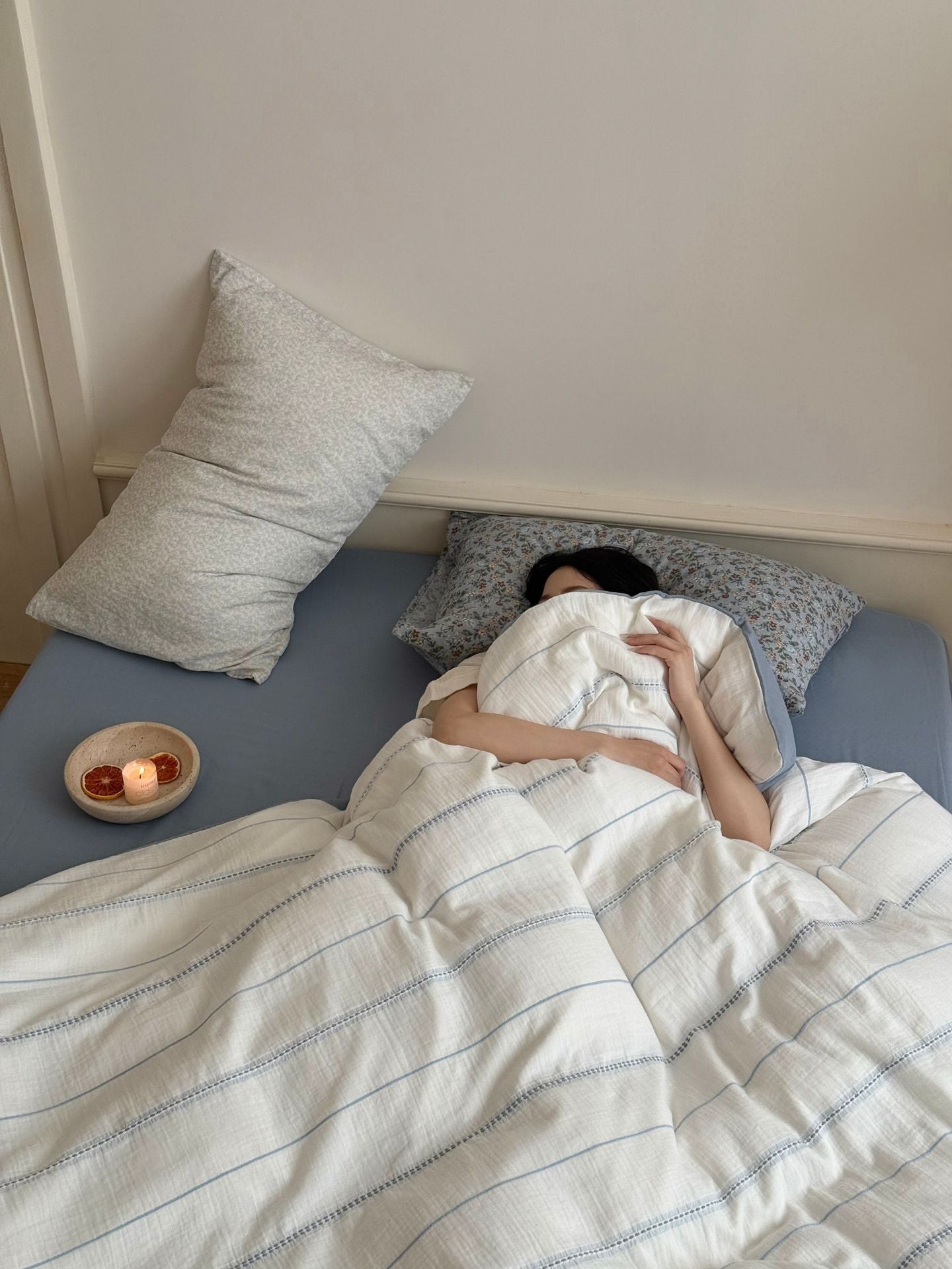 Person lying in bed under a striped duvet with pillows and a small bowl on the nightstand.