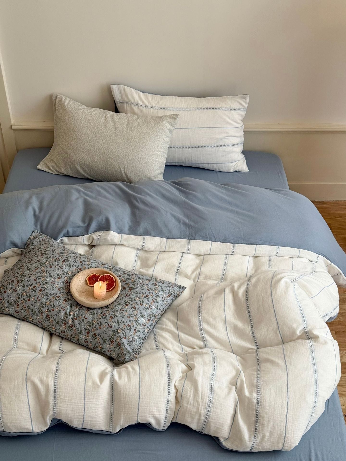Bed with blue sheets, white and gray pillows, and a blanket with a small plate of fruit on a floral pillow.