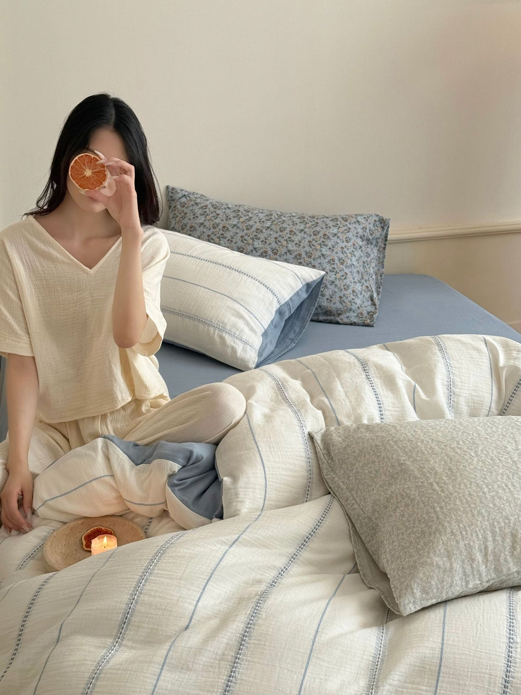 Woman sitting on a bed with pillows and blankets, holding an orange to her eye.