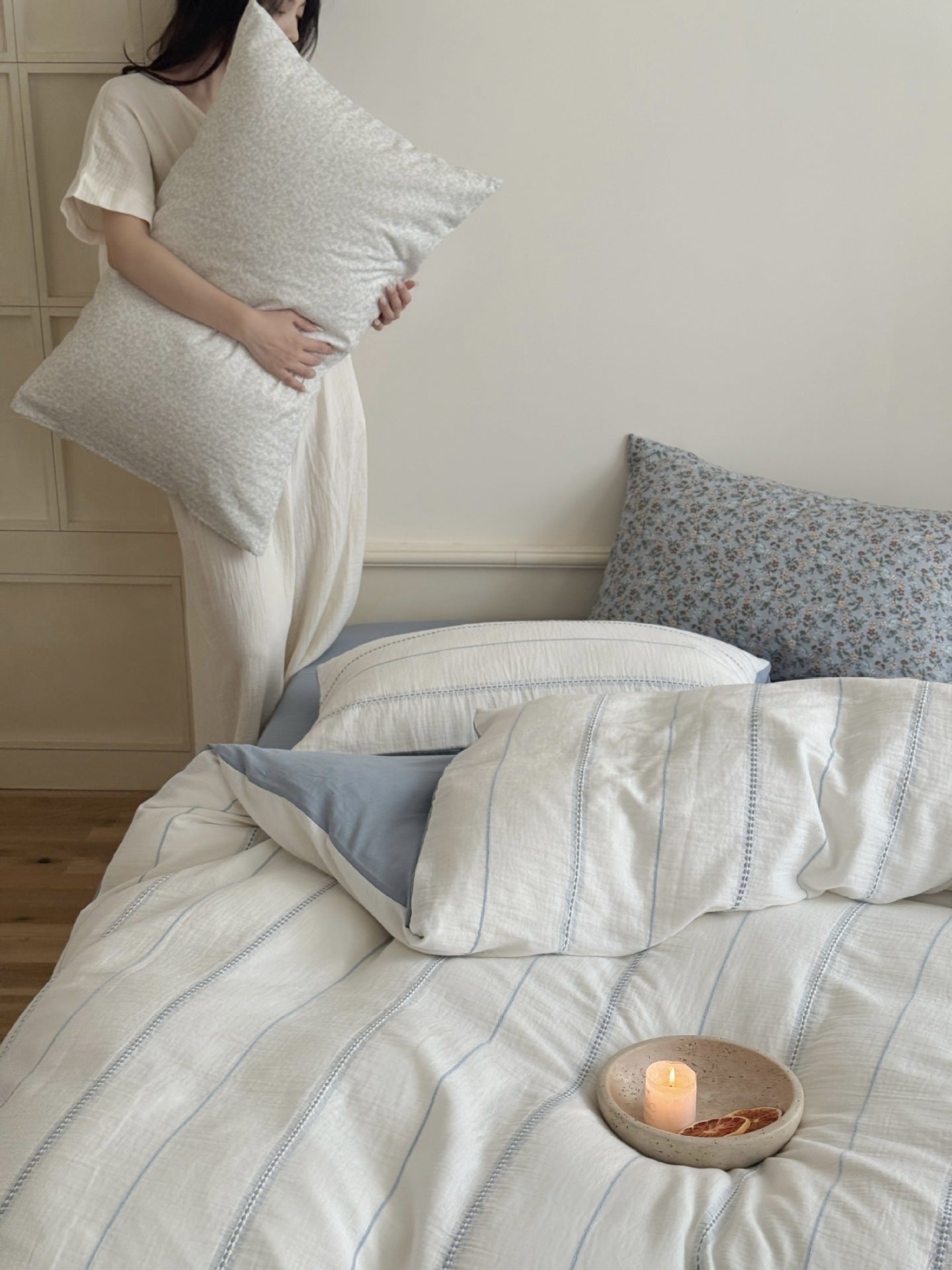 Person holding a pillow next to a bed with striped bedding and a candle.