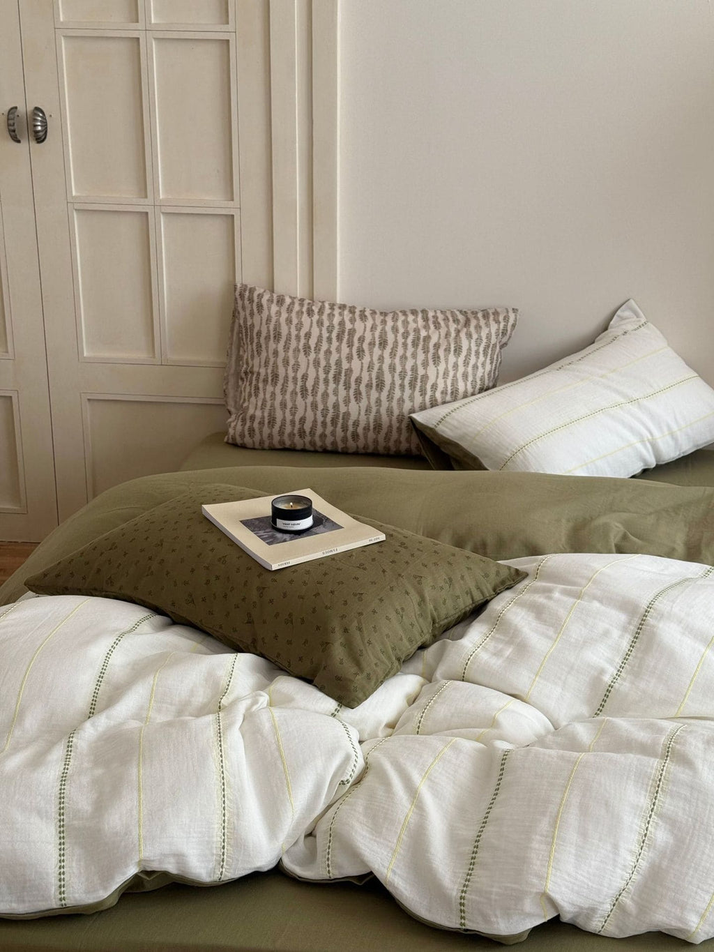 Bed with green and white bedding, pillows, and a small tray with items on a neutral-colored wall.